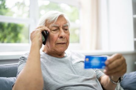 Elderly man on phone looking at a credit card with a concerned expression, sits in a bright room.