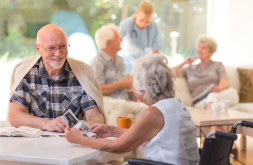 Elderly man and woman enjoy conversation at a senior living facility, with others in the background.
