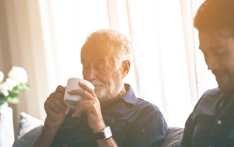 Elderly man drinks coffee while sitting on a sofa next to a younger man in soft natural light.