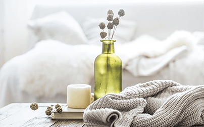 Cozy bedroom scene with a green vase, white candle, and knitted blanket on a wooden table.