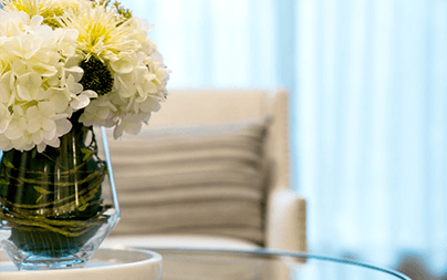 A vase of white flowers sits on a glass table in a bright, modern room with sheer curtains.