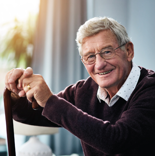 Elderly man with glasses holding a cane, smiling while sitting in a well-lit room