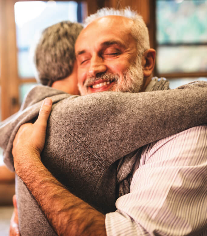 Two men warmly hugging indoors, expressing joy and contentment on their faces.