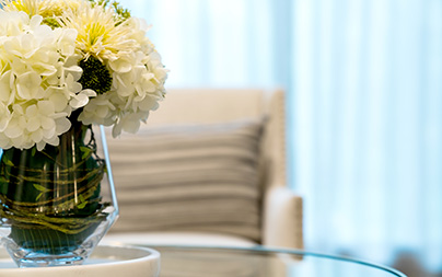 White hydrangeas in a clear glass vase with greenery on a glass table in a cozy living room.